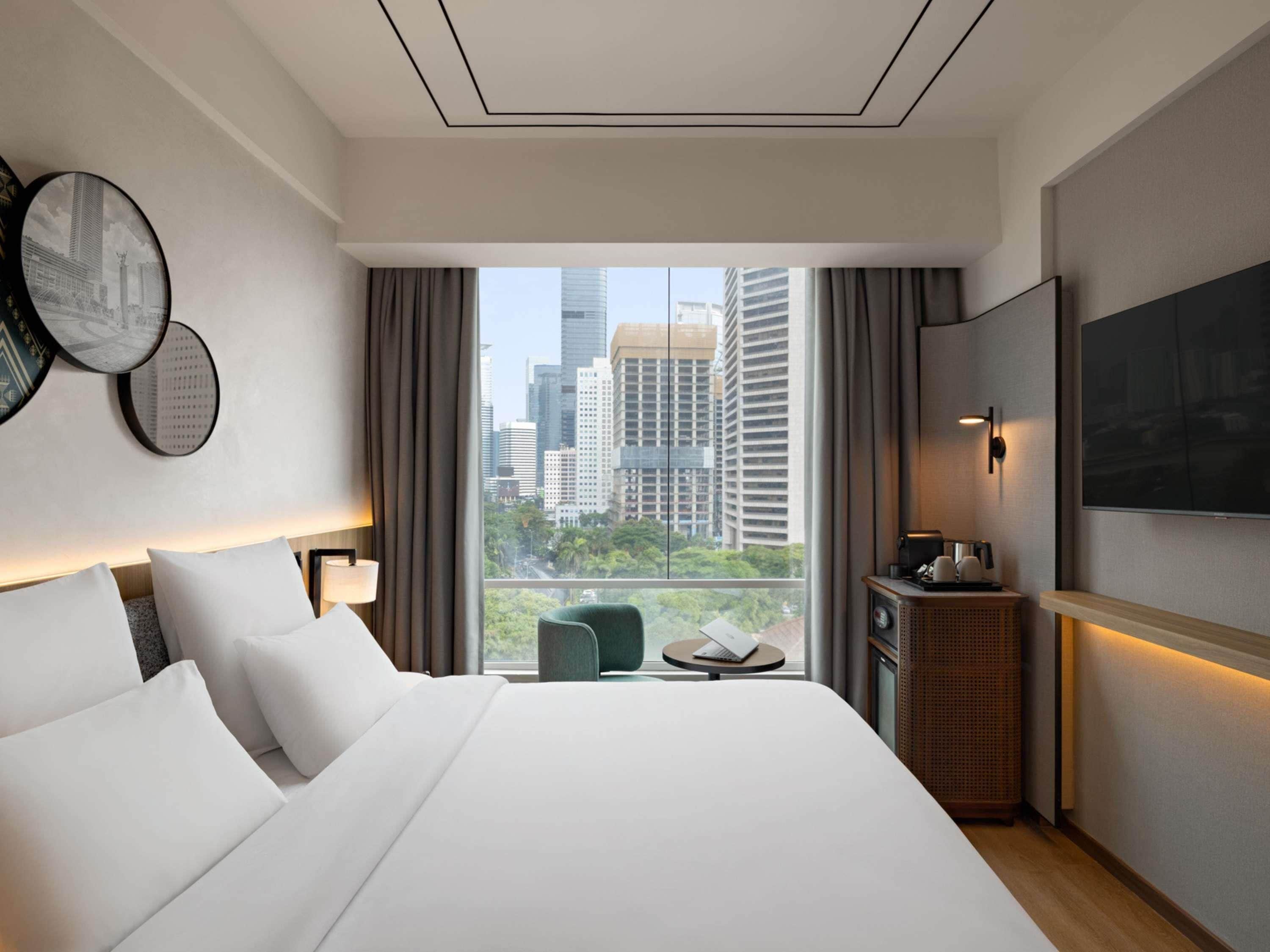 Ein Hotelzimmer mit einem großen Bett, einem Schreibtisch mit Stuhl, einem Fernseher und Blick auf Stadtgebäude.