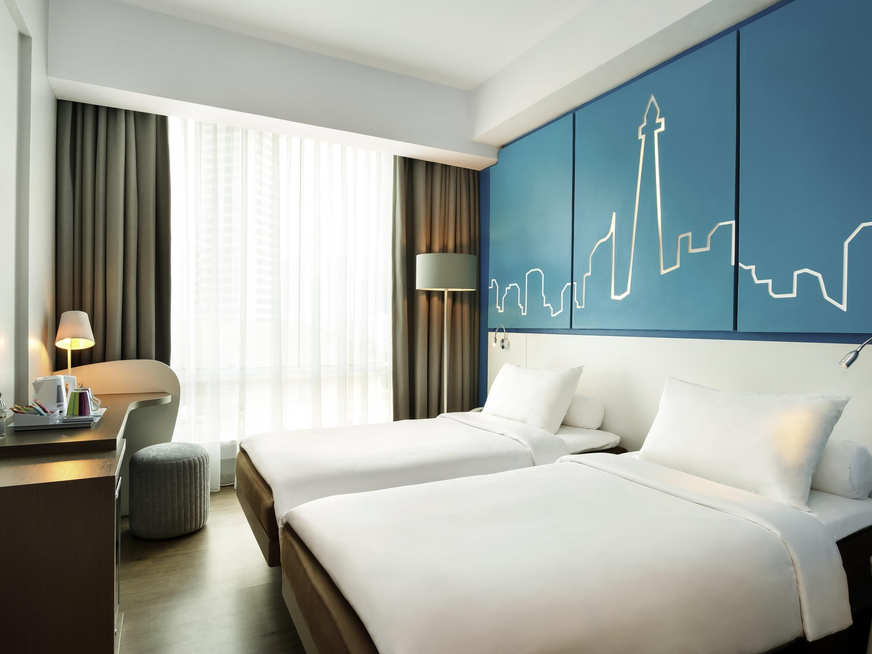 A hotel room with two beds, a desk with a lamp, a chair, and a large window with curtains.