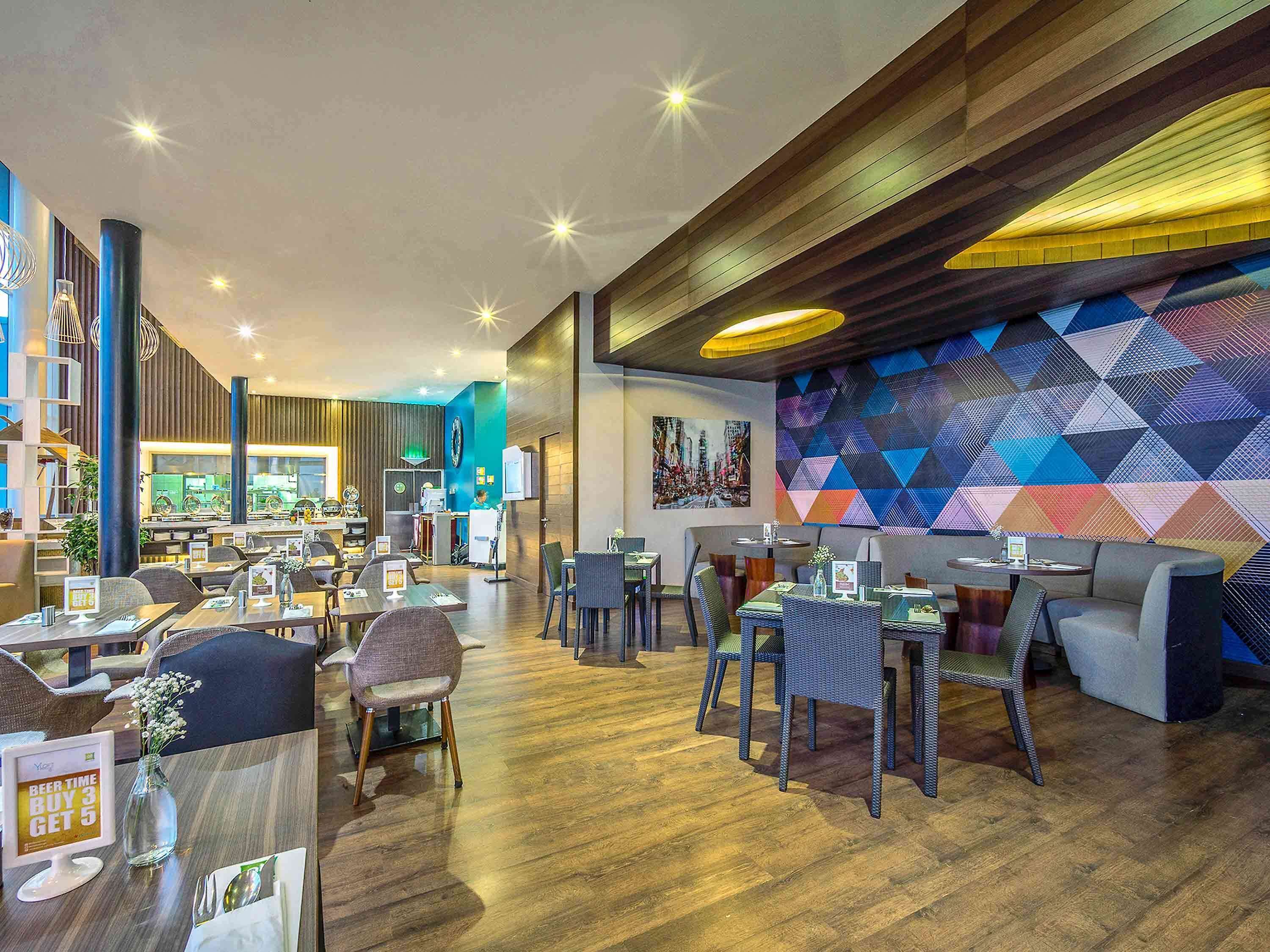 Un restaurante moderno con suelos de madera, mesas y sillas altas. Se observa un vibrante arte mural geométrico y un reloj grande en la pared.