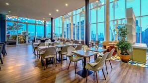 Un restaurant spacieux avec de hauts plafonds et de grandes fenêtres offrant une vue sur la ville. Les tables sont disposées pour accueillir les convives, et des plantes en pot apportent une touche de verdure.