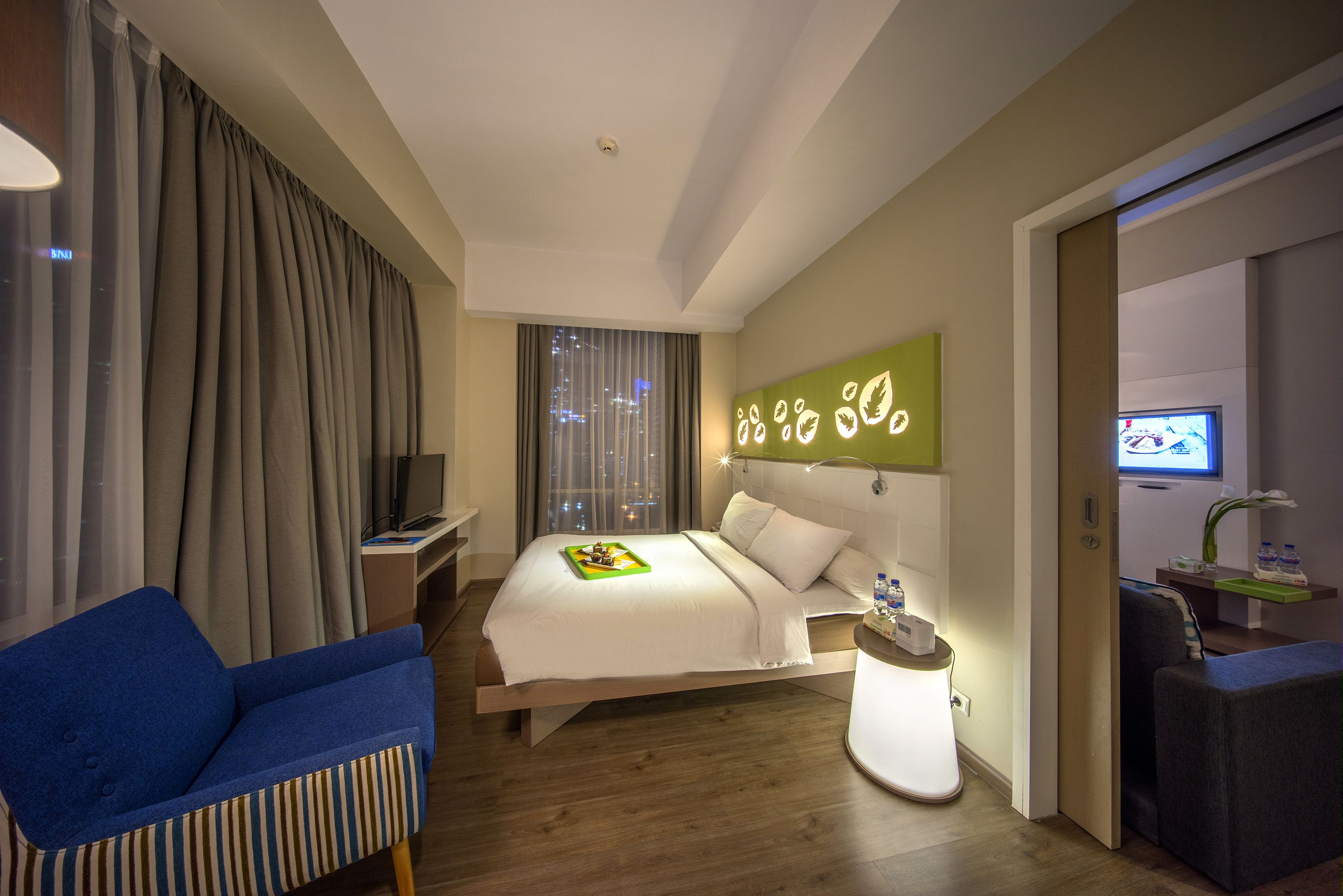 Une chambre d’hôtel moderne avec un lit, un fauteuil bleu, une table de chevet avec une lampe et un téléviseur posé sur un meuble.