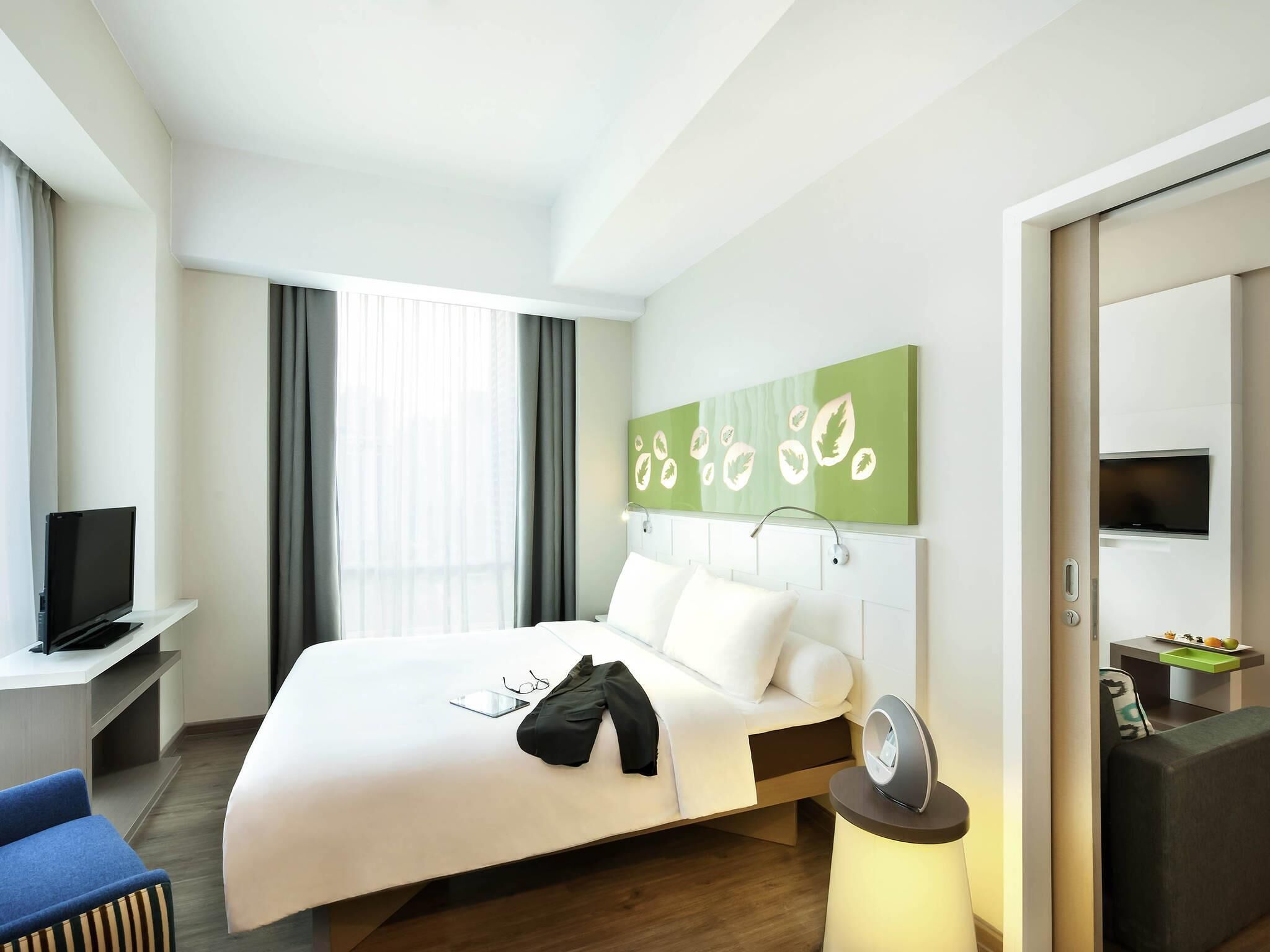 A hotel room with a bed, a desk with a TV, a bedside table with a lamp, and a wall-mounted artwork.