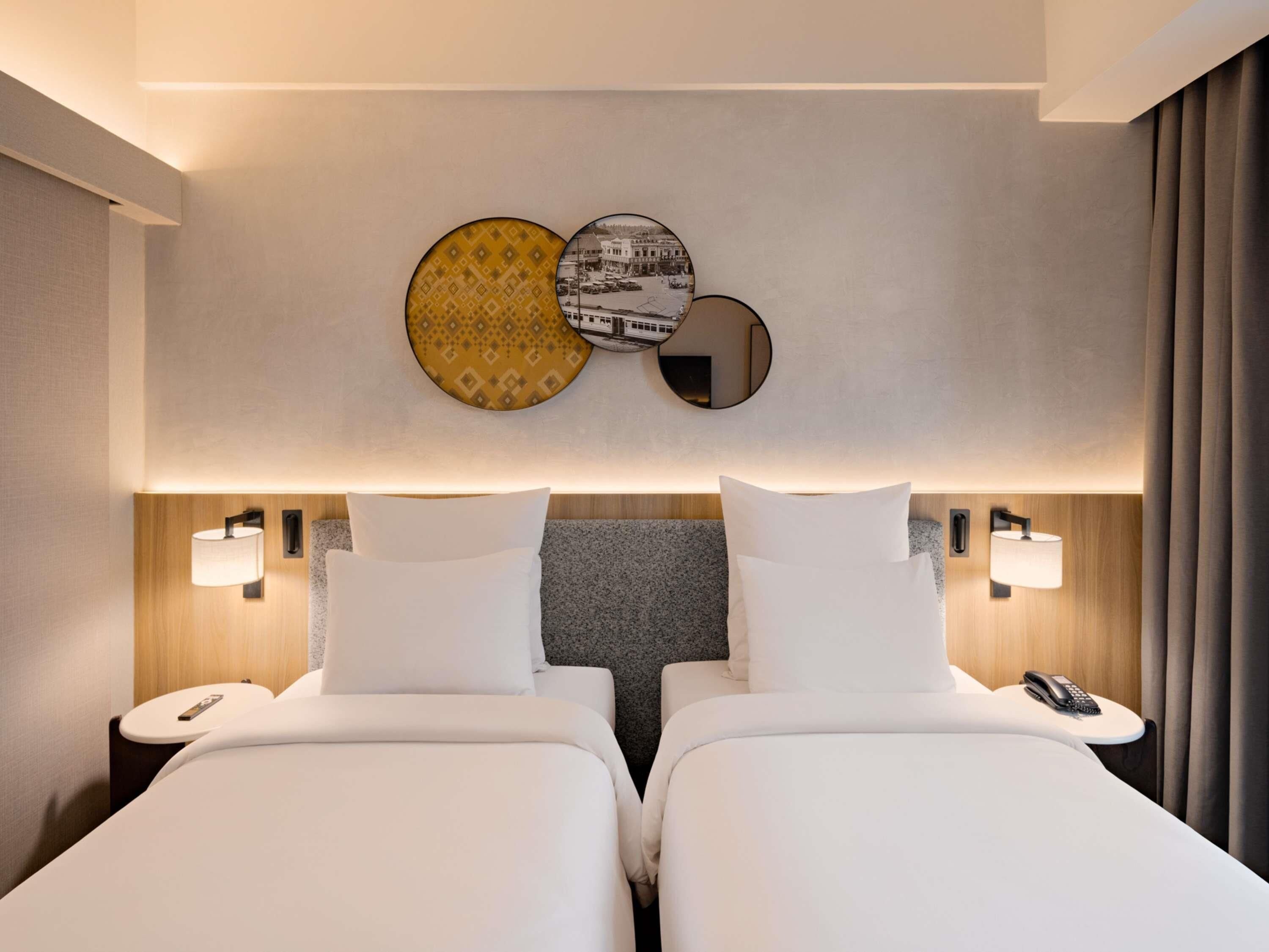 Habitación de hotel con dos camas, cabeceros, lámparas de noche, un teléfono y obras de arte colgadas en la pared.