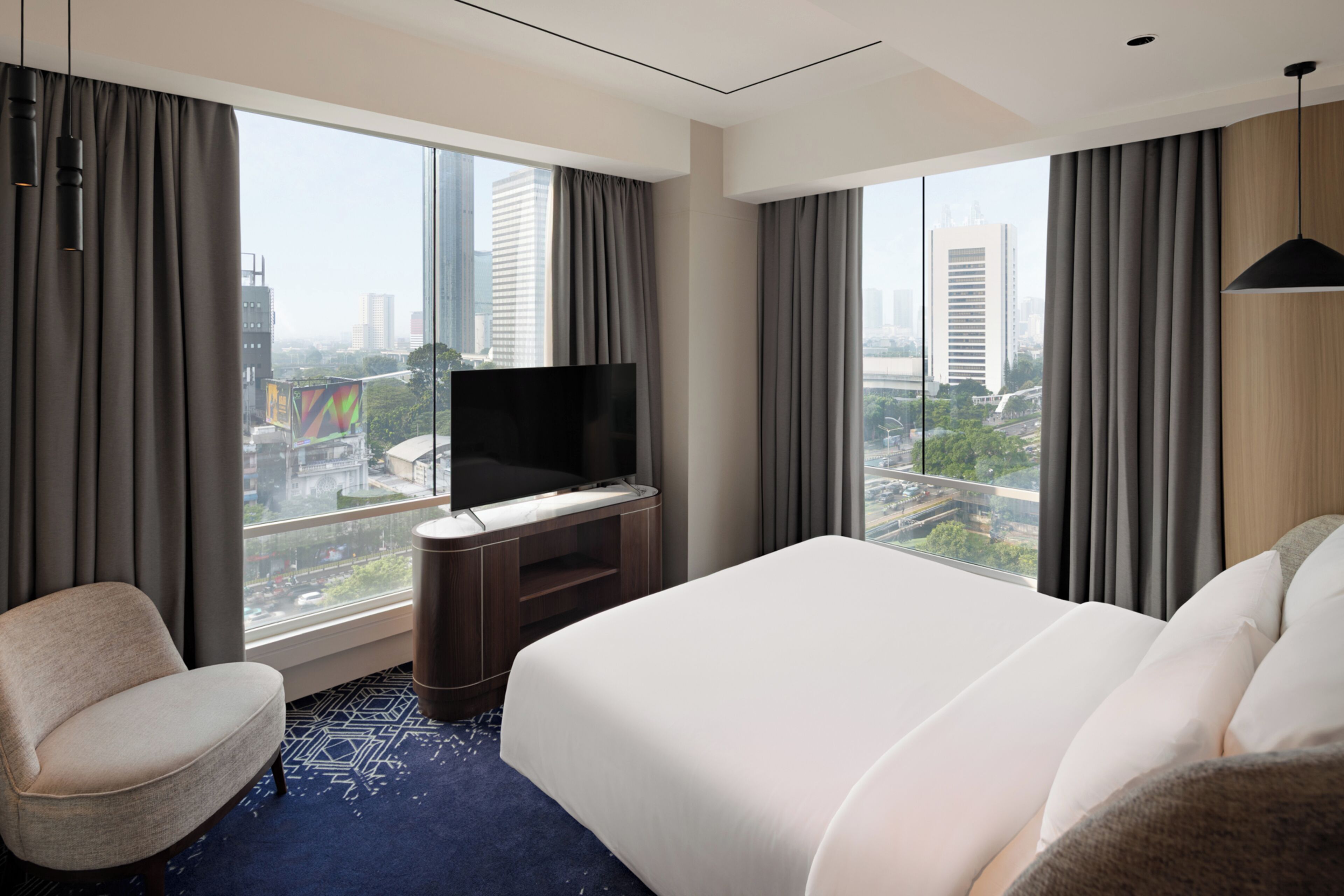 A hotel room with a bed, a TV, a chair, and large windows with city views.
