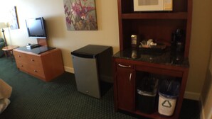 A hotel room with a television, a small refrigerator, a microwave on a wooden cabinet, a trash bin, and a painting on the wall.