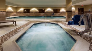 Une piscine intérieure avec un jacuzzi au centre, entourée de chaises longues et d’une chaise de maître-nageur.