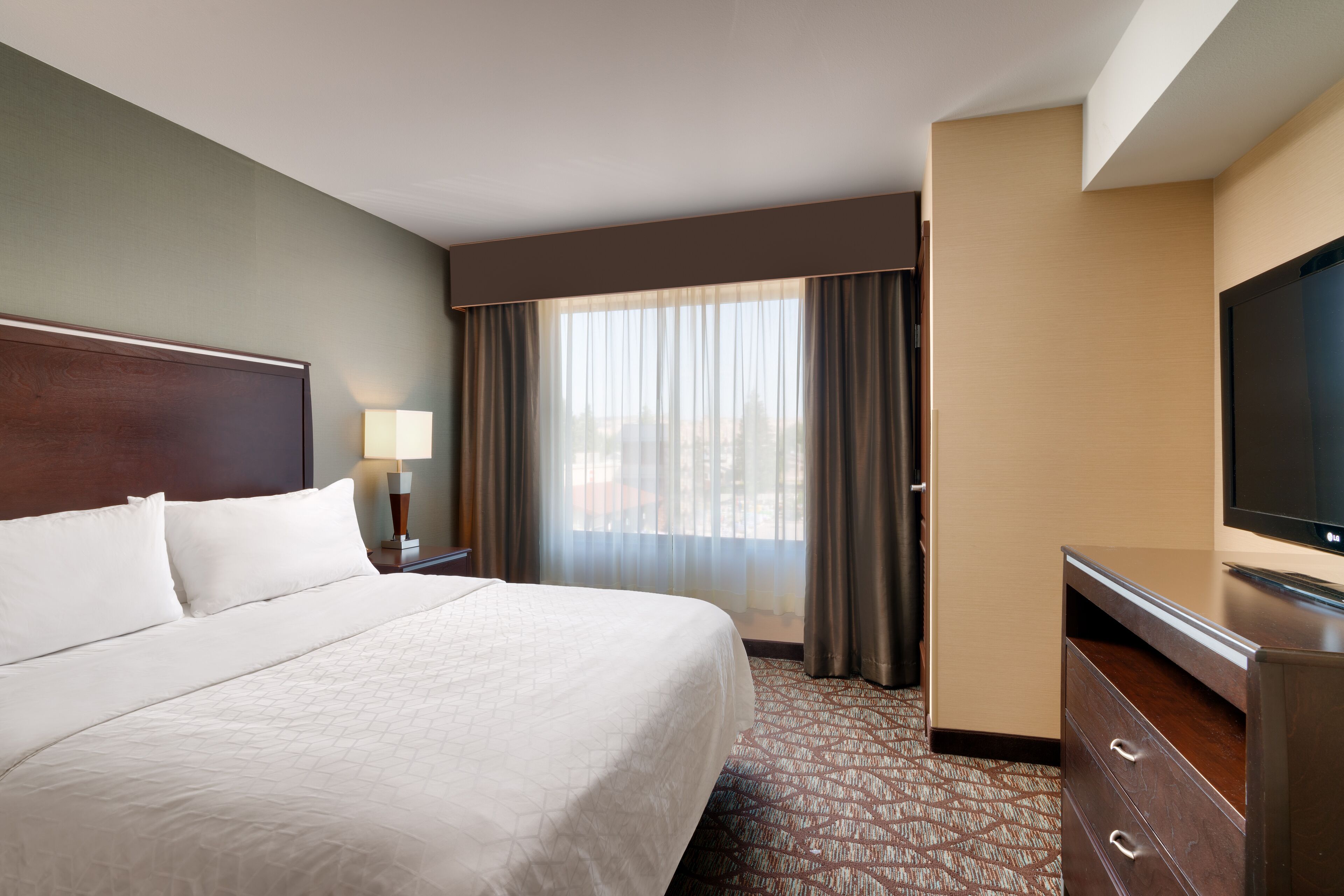 A hotel room with a large bed, a TV on a dresser, a window with curtains, and a lamp on a nightstand.