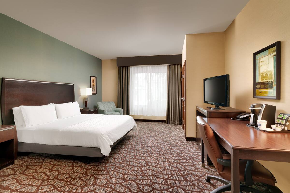 A hotel room with a large bed, a desk with a computer, a television, and a window with curtains.