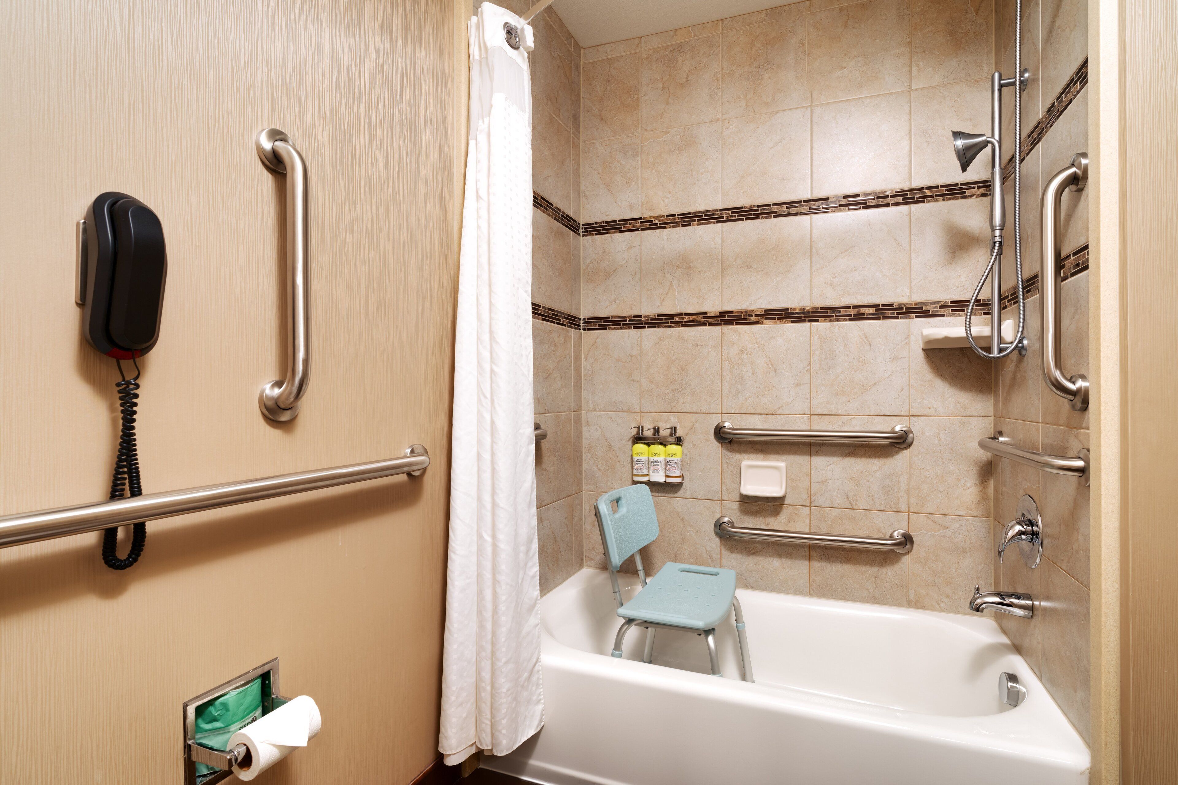 A bathroom with a bathtub, grab bars, a shower chair, a toilet paper holder, and a telephone.