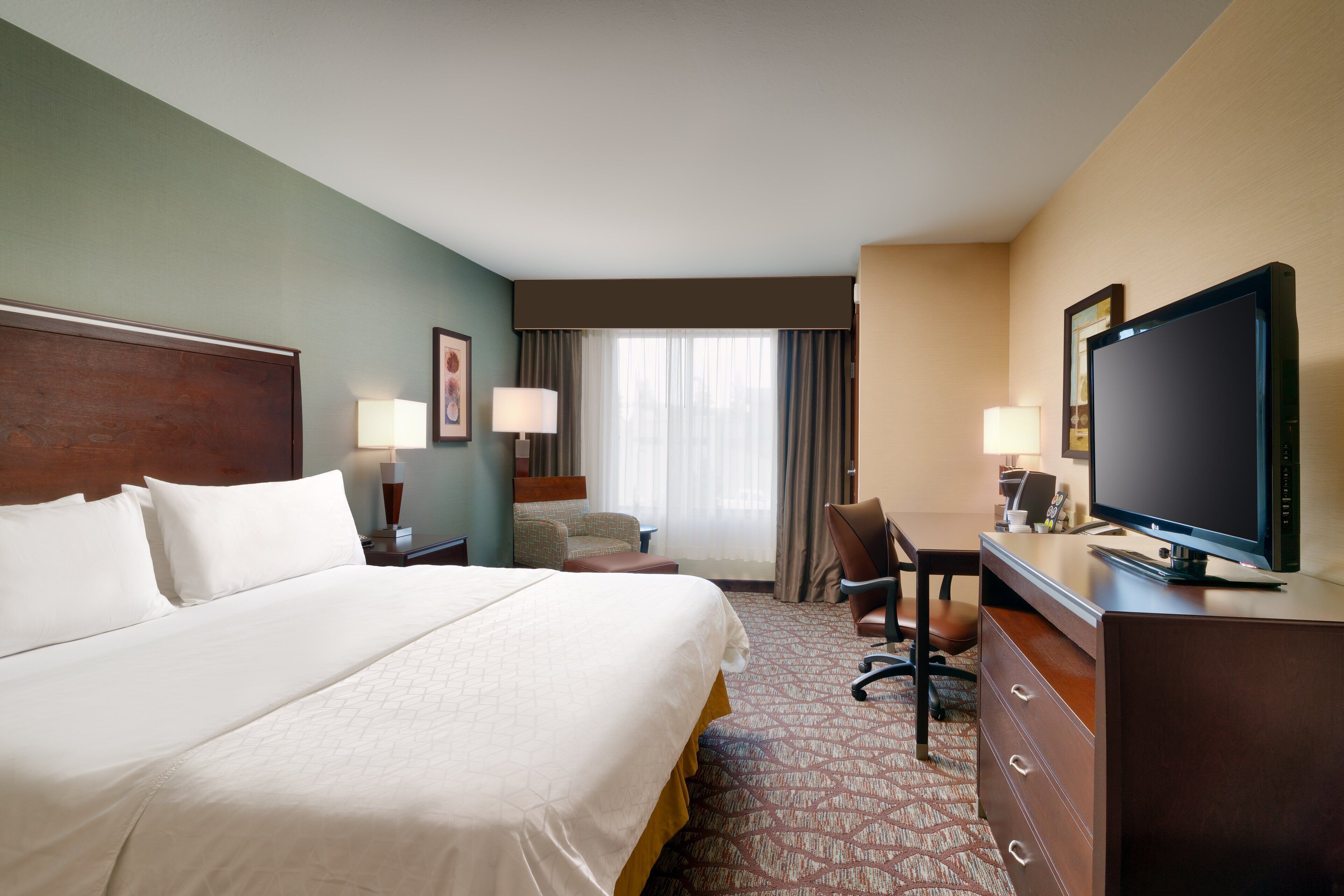 Quarto de hotel com uma cama grande, uma escrivaninha com televisão, uma cadeira e uma luminária de mesa.