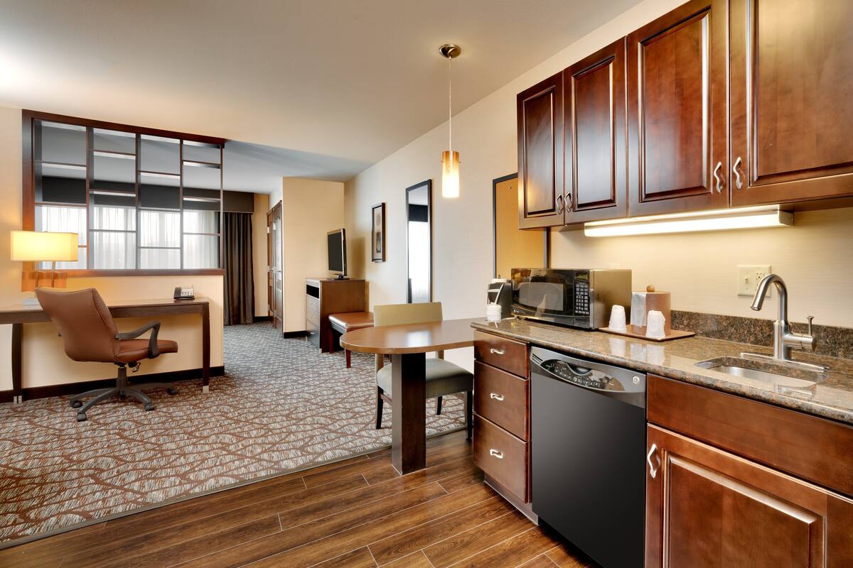 A hotel room with a kitchenette, a dining area with a table and chairs, a desk with a chair, and a large mirror.
