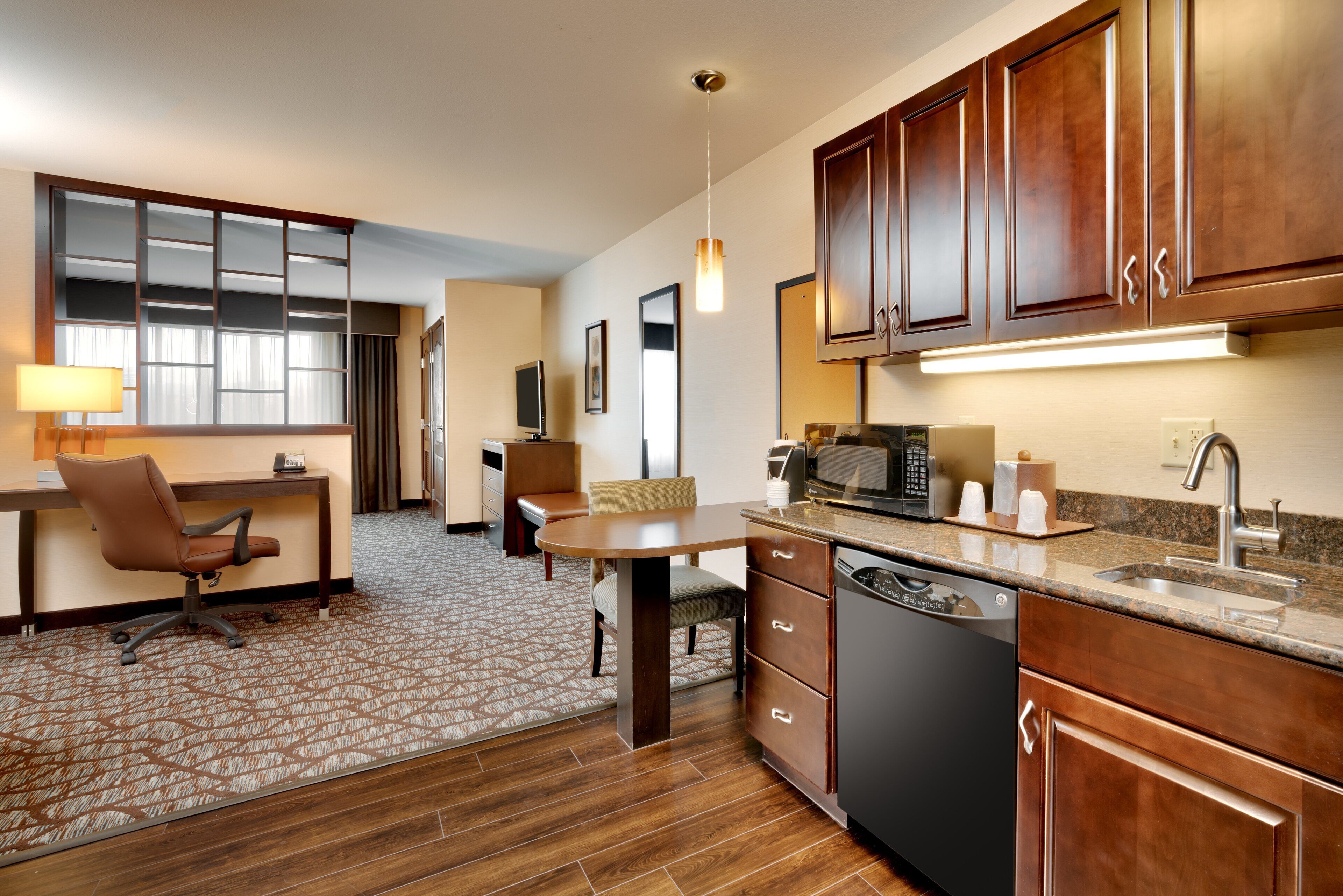 A hotel room with a kitchenette, a desk with a chair, a dining table with chairs, and a patterned carpet.