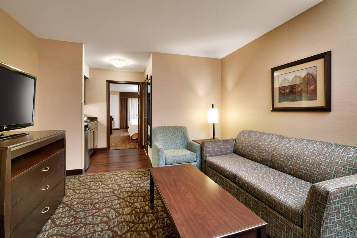 A hotel room with a sofa, armchair, TV, and a painting on the wall.