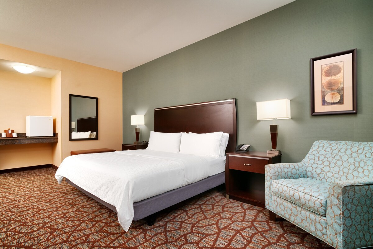 Une chambre d’hôtel avec un lit, des tables de chevet, une chaise, un miroir et un tableau encadré au mur.