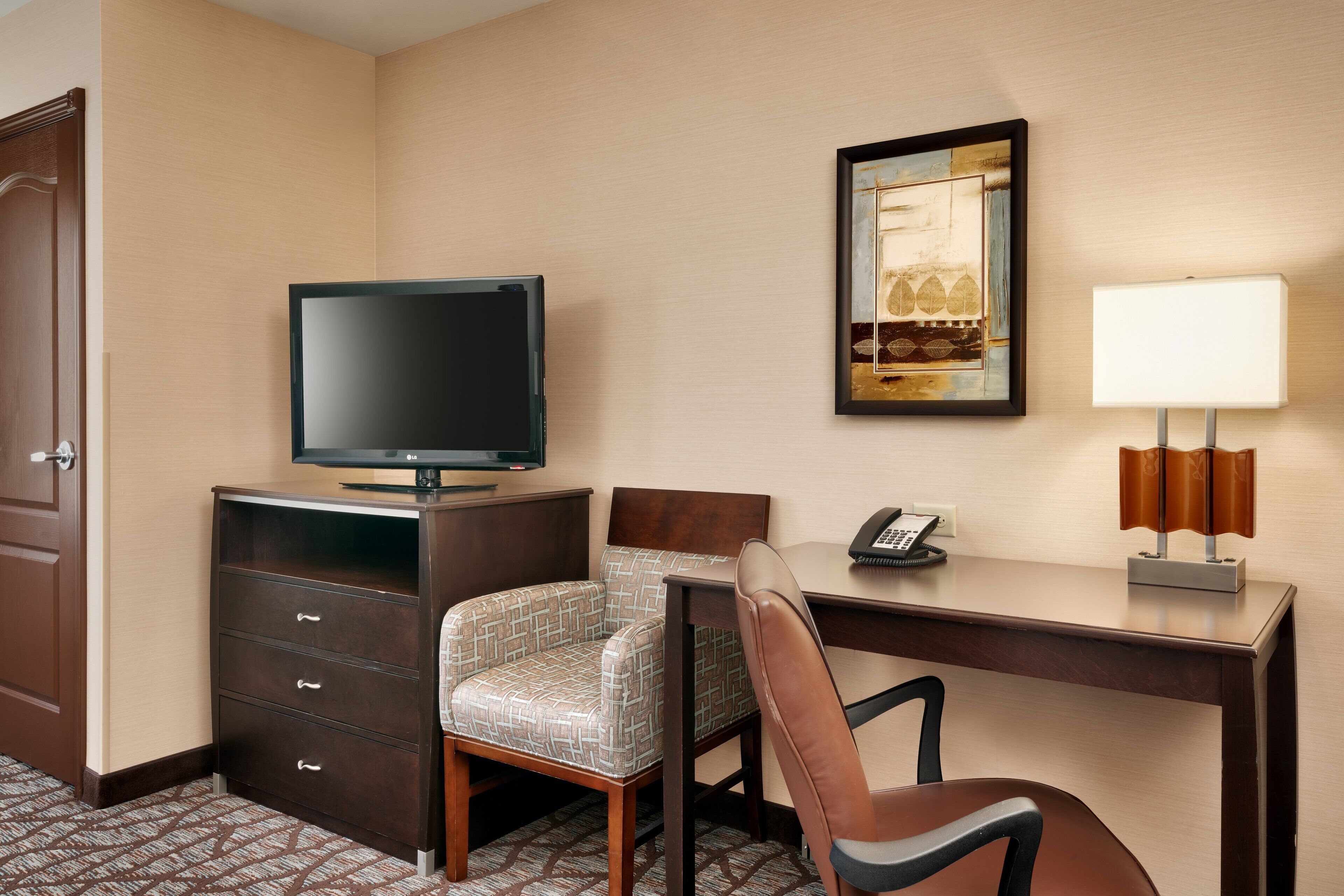 Habitación de hotel con televisor de pantalla plana, escritorio con lámpara, teléfono, una silla estampada y un armario.