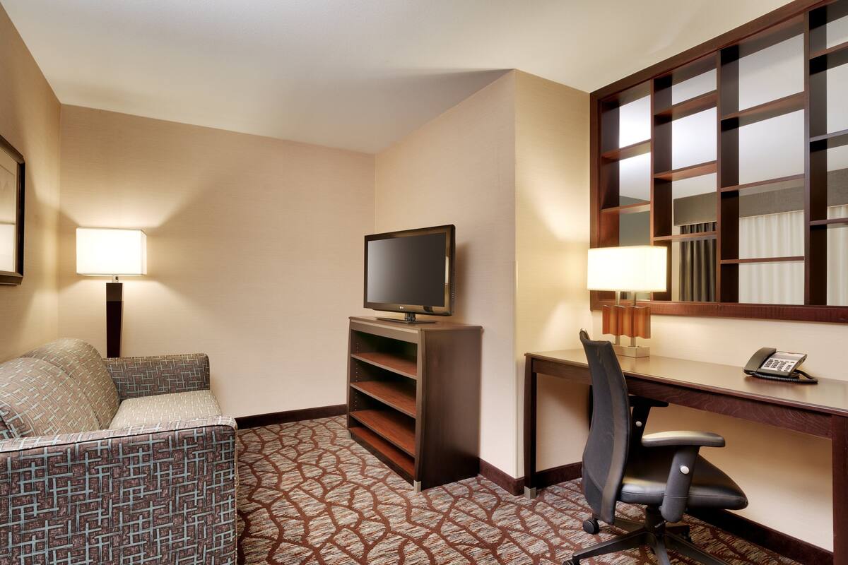 A hotel room with a sofa, a desk with a computer and phone, a TV on a stand, and a patterned carpet.