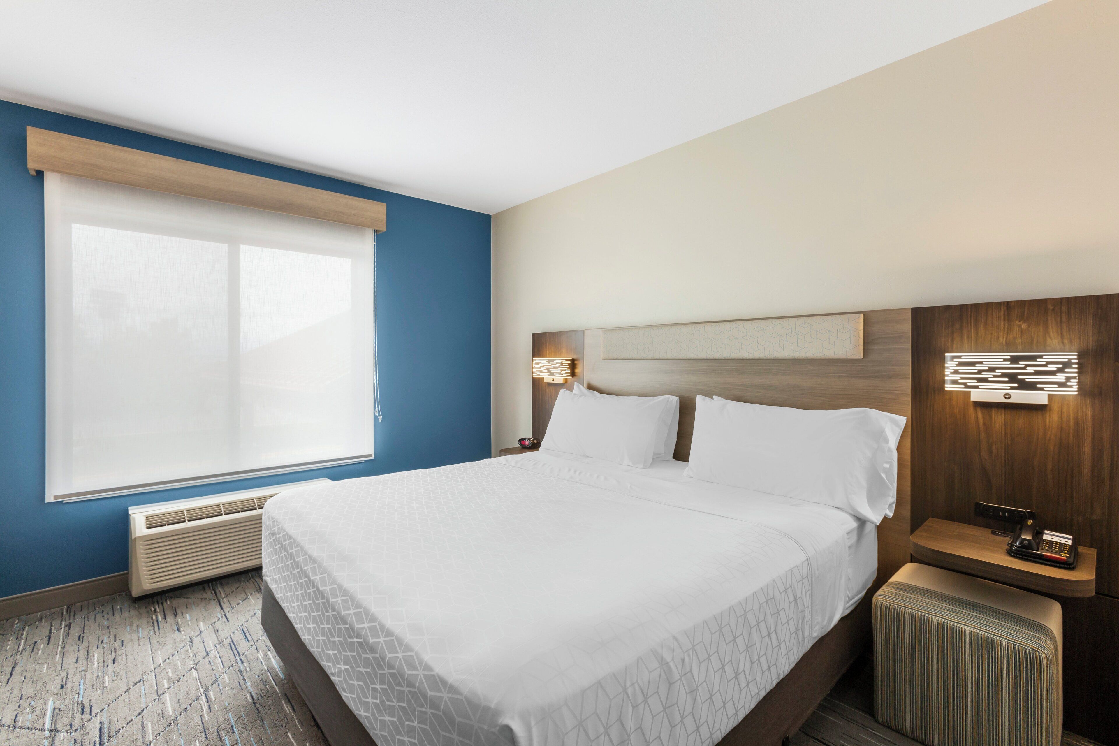 Een hotelkamer met een groot bed, een raam met luxaflex, een nachtkastje met een lamp en een wandlamp.