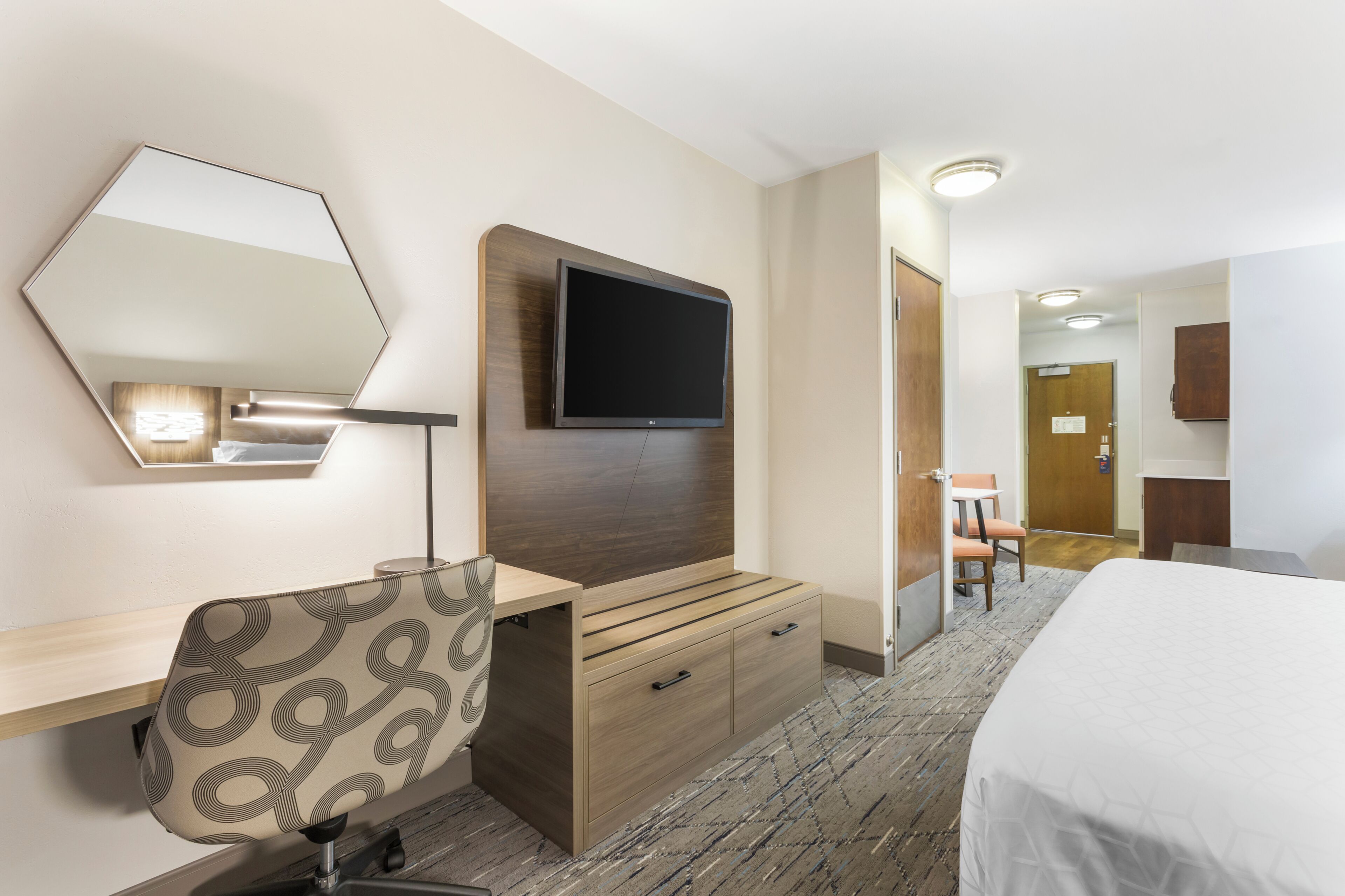 Una habitación de hotel con cama, un escritorio y una silla, un televisor montado en un panel de madera, un espejo y una puerta que da a otra habitación.