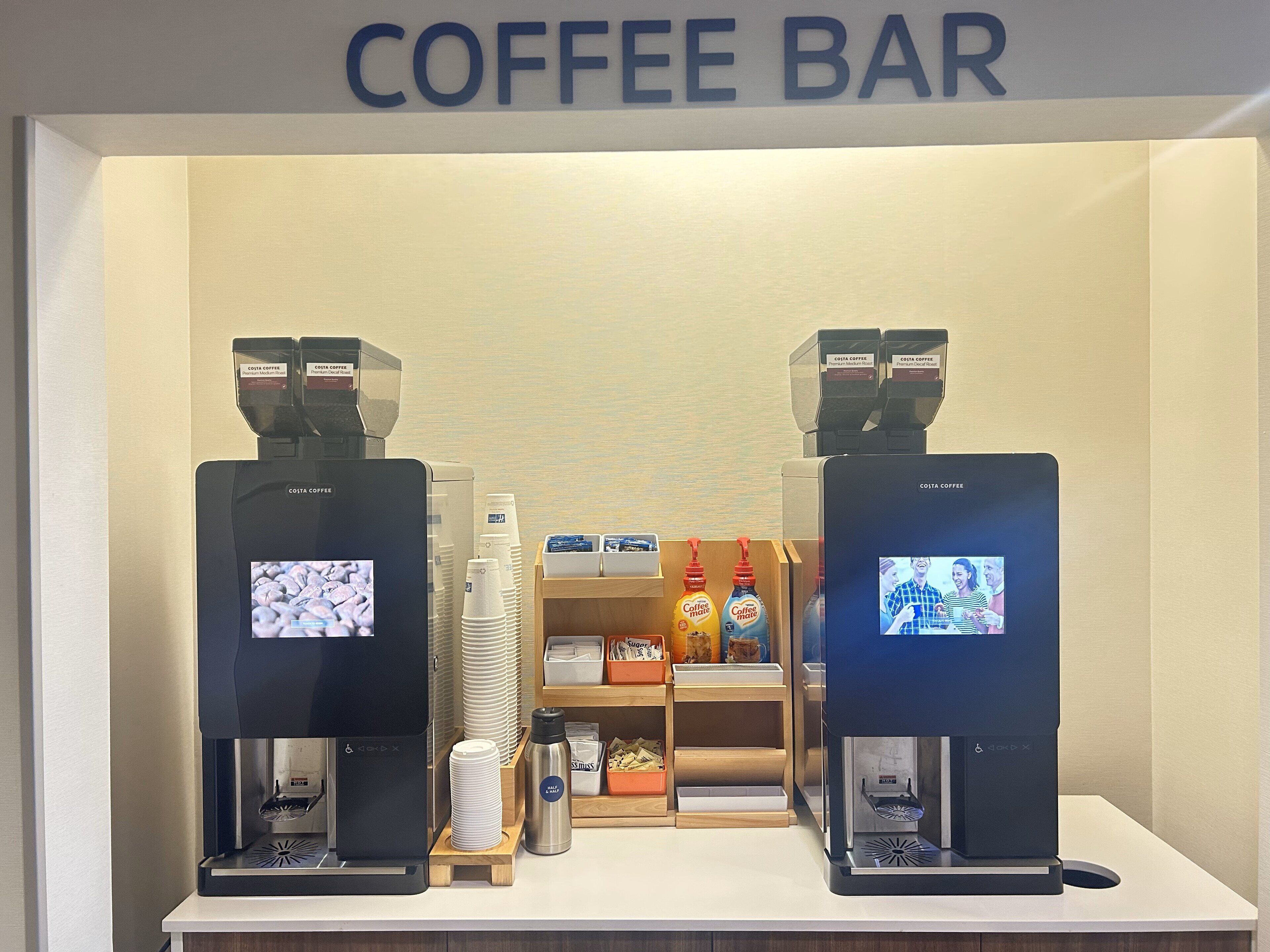 A coffee bar with two coffee machines, disposable cups, and a coffee dispenser.