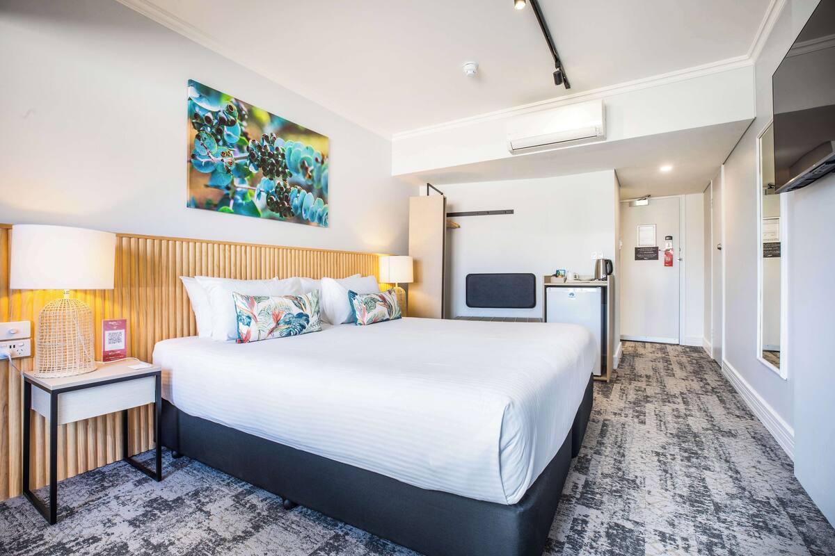 A modern hotel room with a large bed, a bedside table with a lamp, a flat-screen TV, and a small kitchenette.