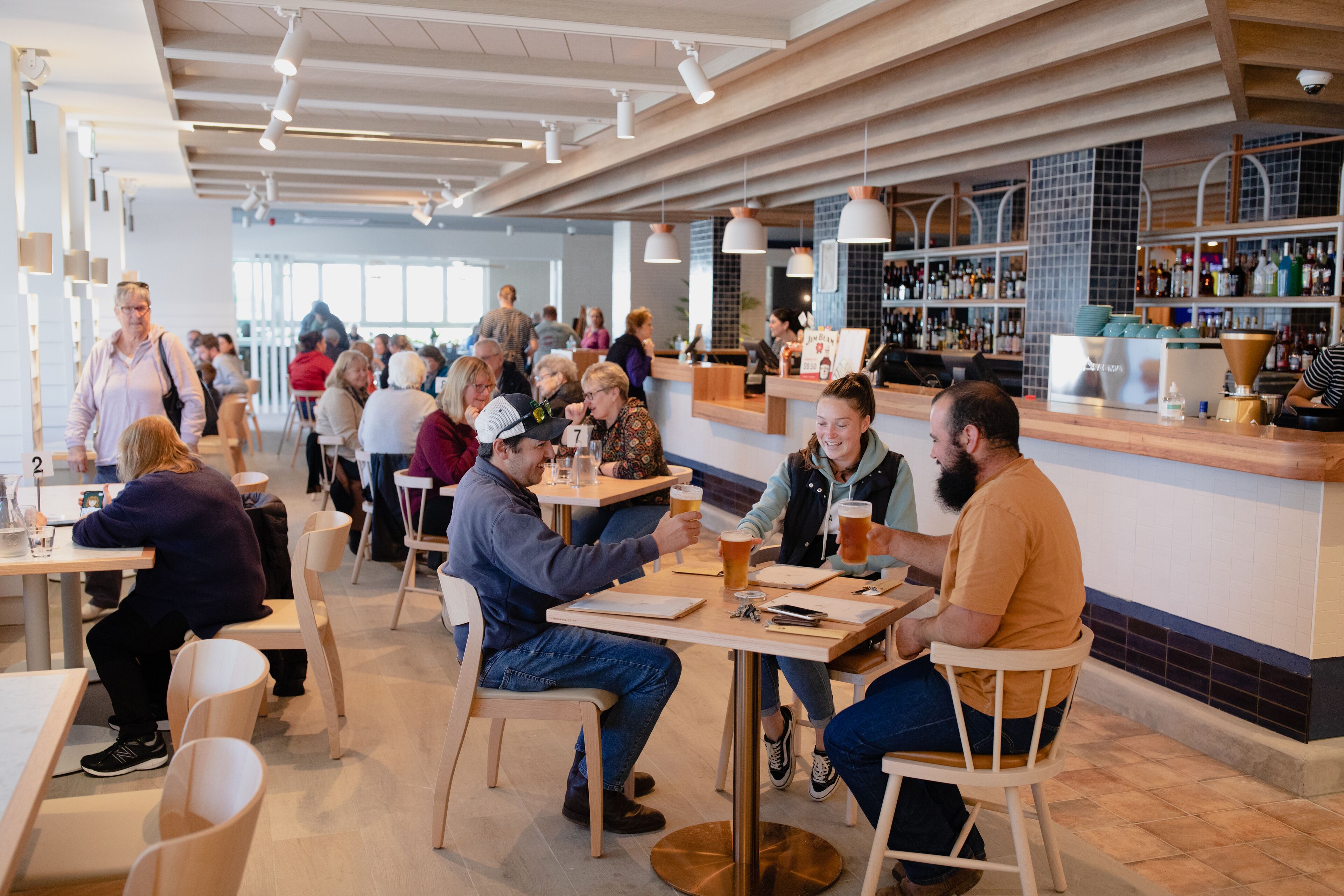 Een ruime, overdekte eethoek met meerdere tafels en stoelen, een bar met diverse flessen en een betegelde achterwand.