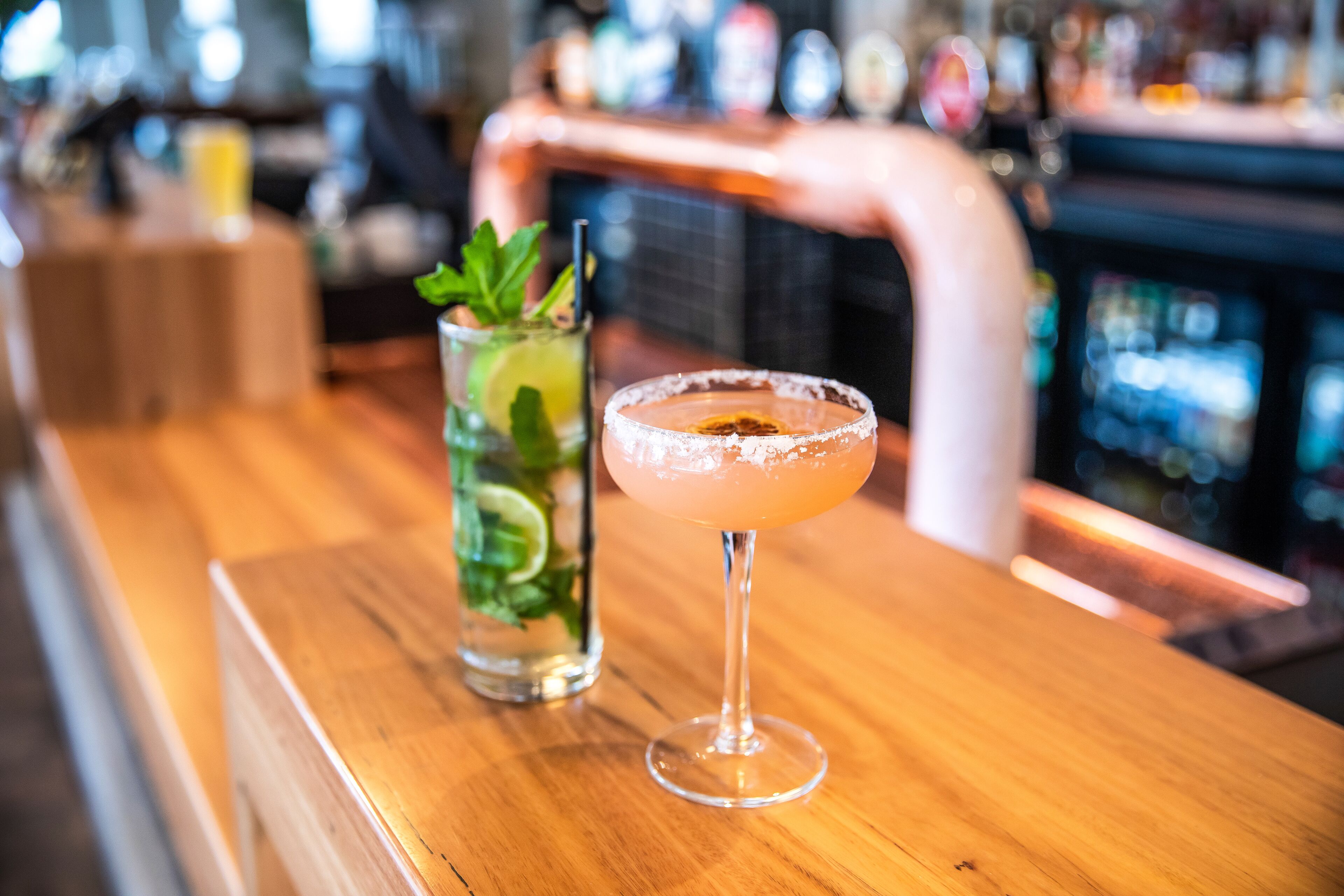 A bar setting with a cocktail and a glass of limeade on a wooden bar counter.
