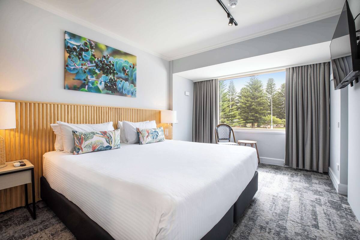 A hotel room with a large bed, a nightstand, a chair, a small table, a painting on the wall, and a window with curtains.