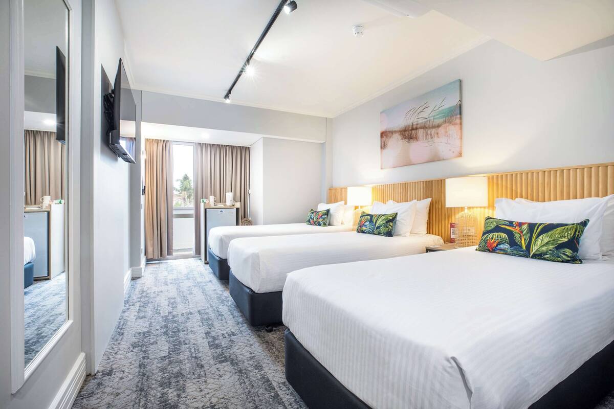 A hotel room with three beds, a television, a painting, and a view of the outdoors.