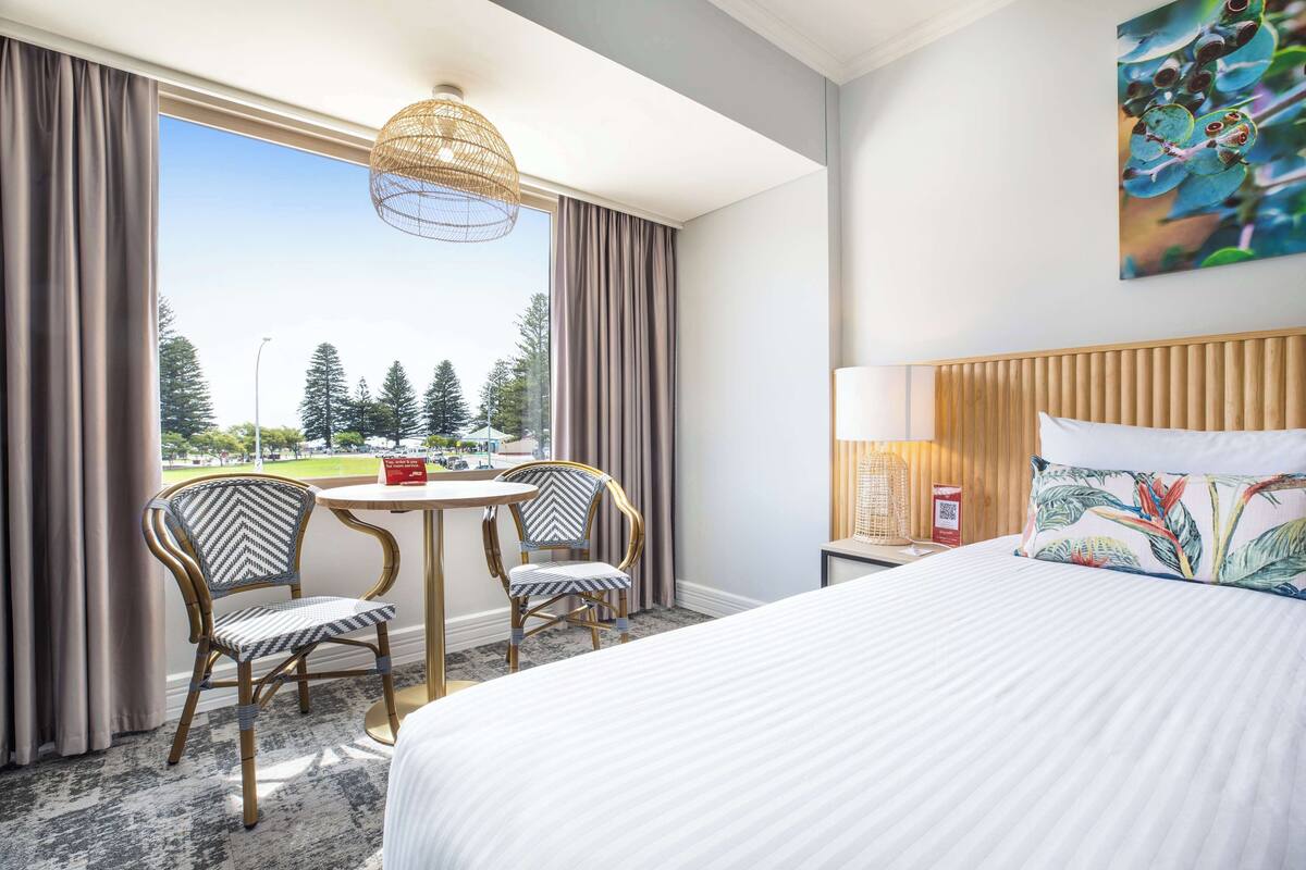 A hotel room with a bed, two chairs, a small table, a lamp, and a view of trees and a parking lot.