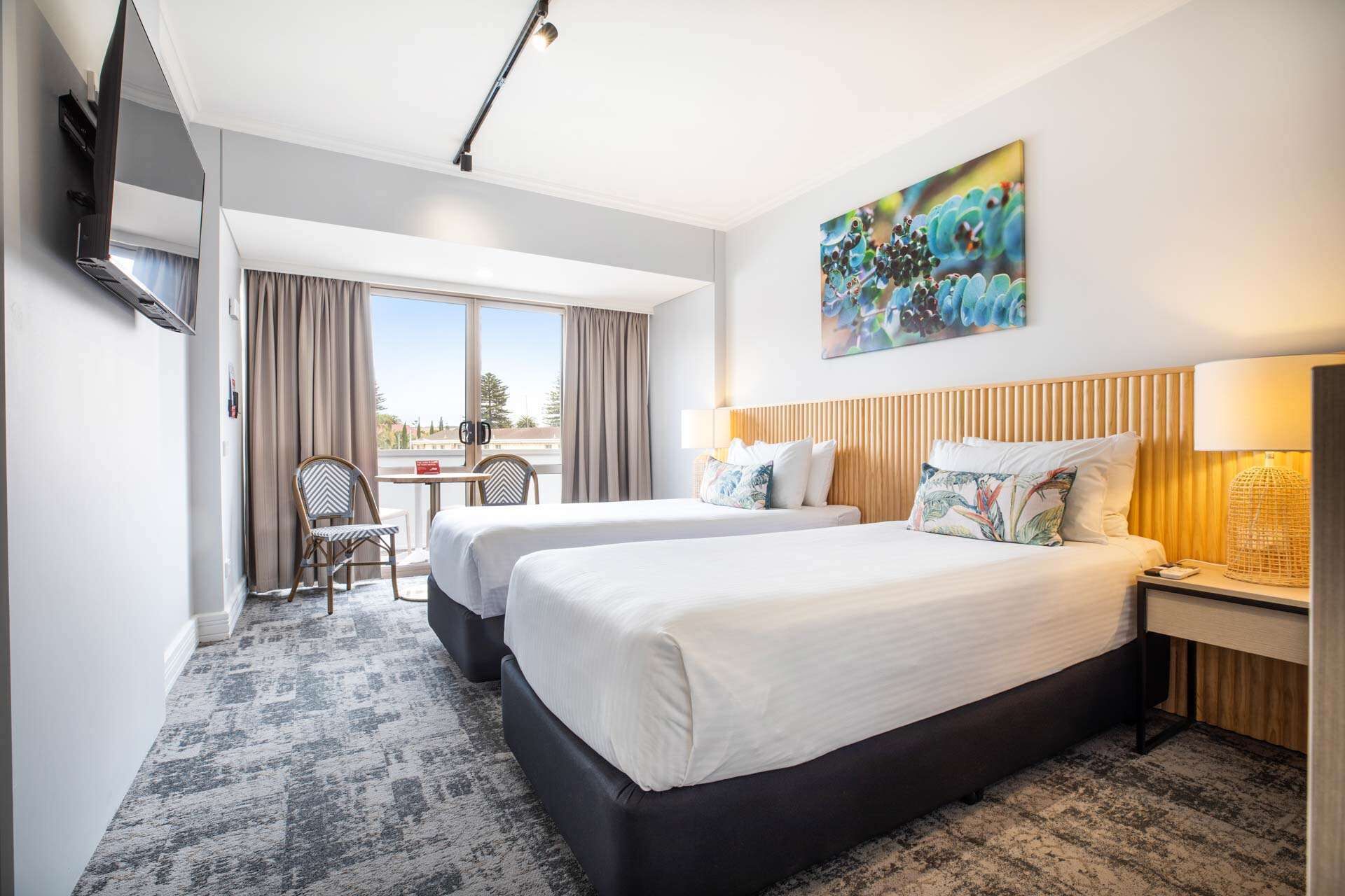 A hotel room with two beds, a desk, and a chair. There is a painting on the wall and a TV mounted on the wall.
