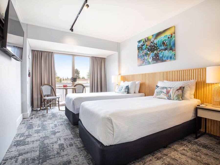 A hotel room with two beds, a desk, and a chair. There is a painting on the wall and a TV mounted on the wall.