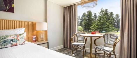 A hotel room with a bed, a table with two chairs, a lamp, and a view of trees through large windows.