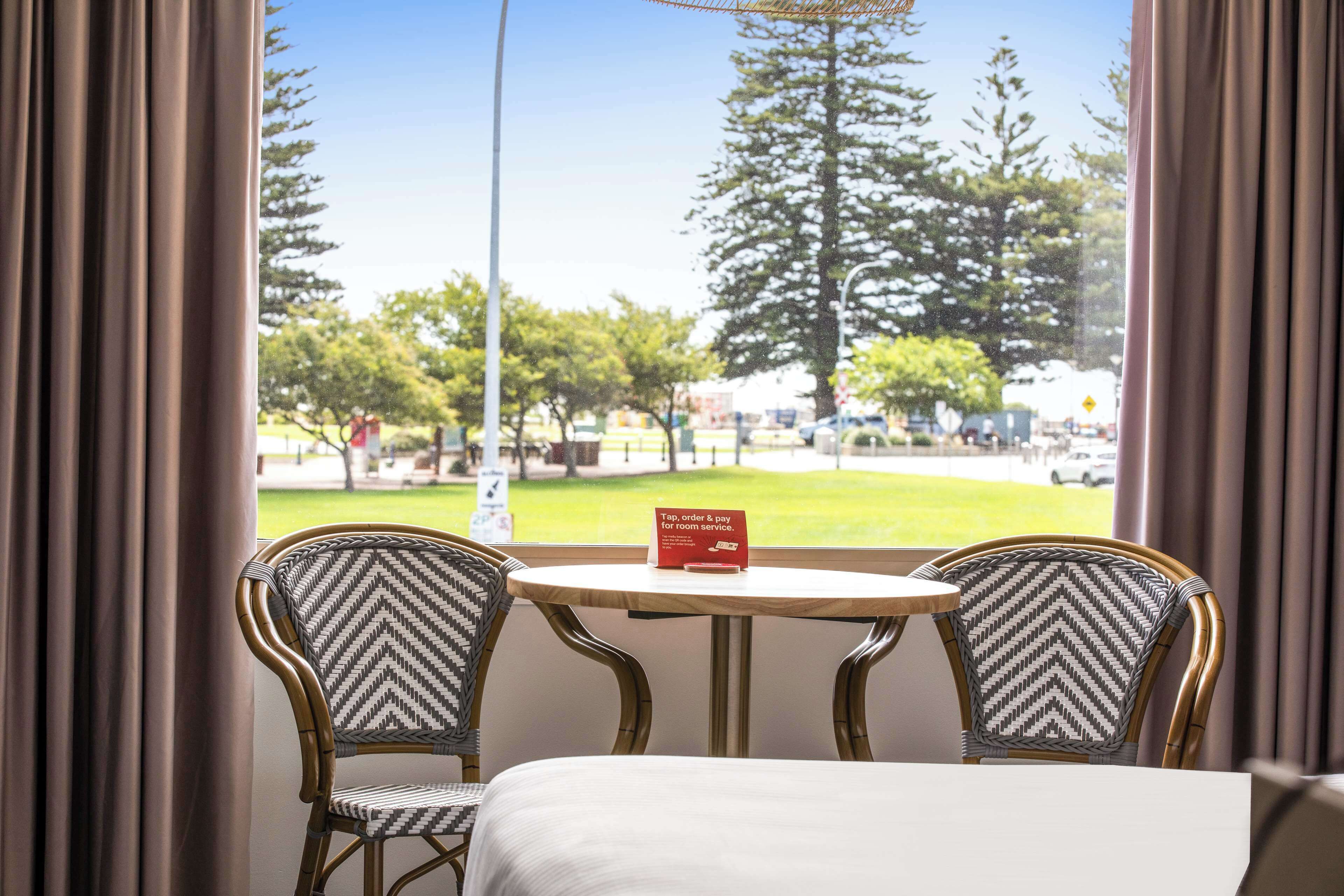 A hotel room with a bed, two chairs, a small table, and a view of a park through large windows.