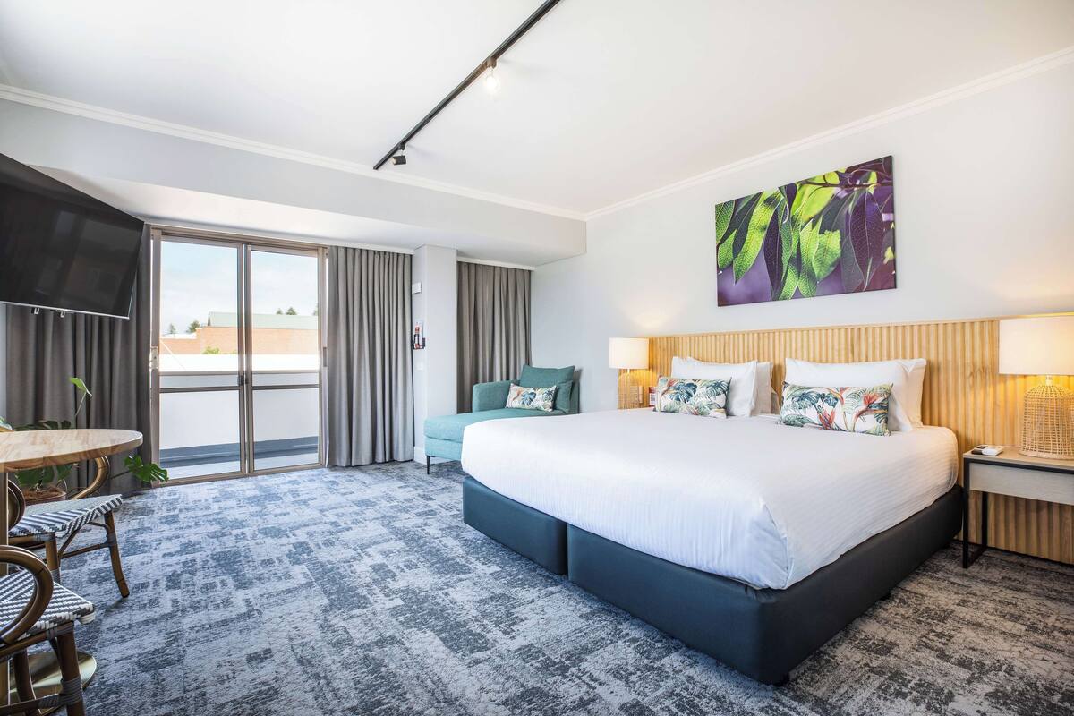 A hotel room with a large bed, a dining table, a TV, and a balcony with curtains.