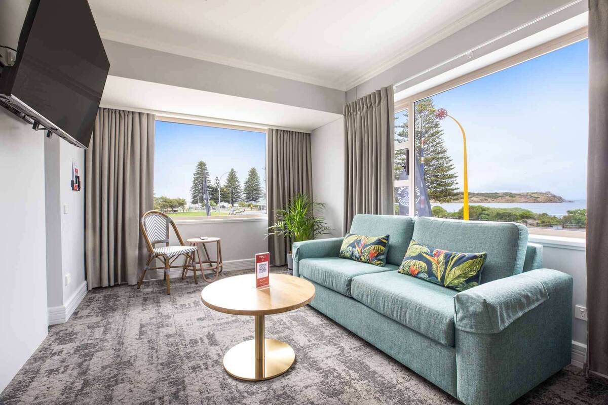 A room with a sofa, a chair, a small table, and large windows offering a view of the sea and trees.