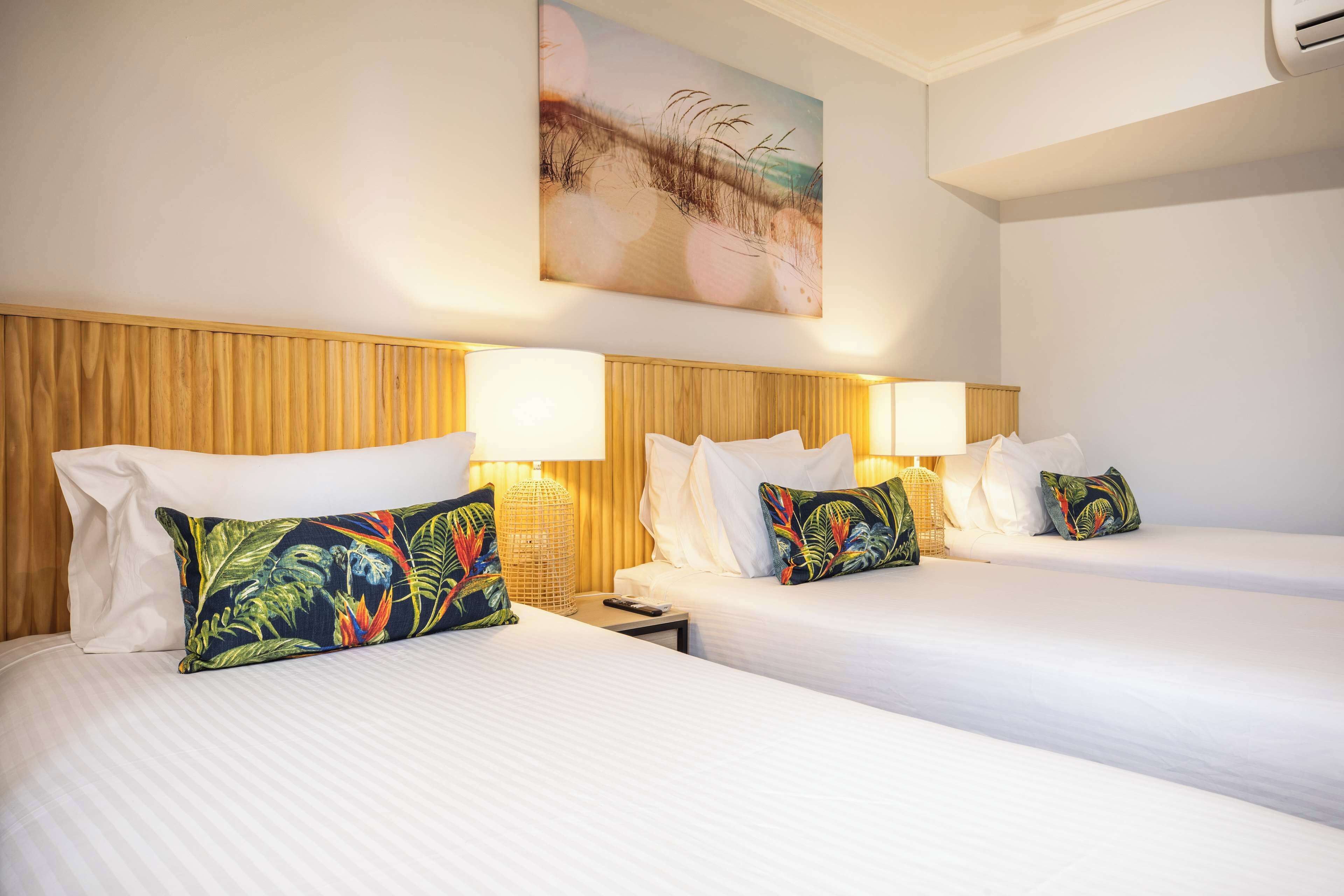 A hotel room with two beds, each with white linens and tropical-themed pillows. There are wall-mounted lamps and a large painting above the beds.