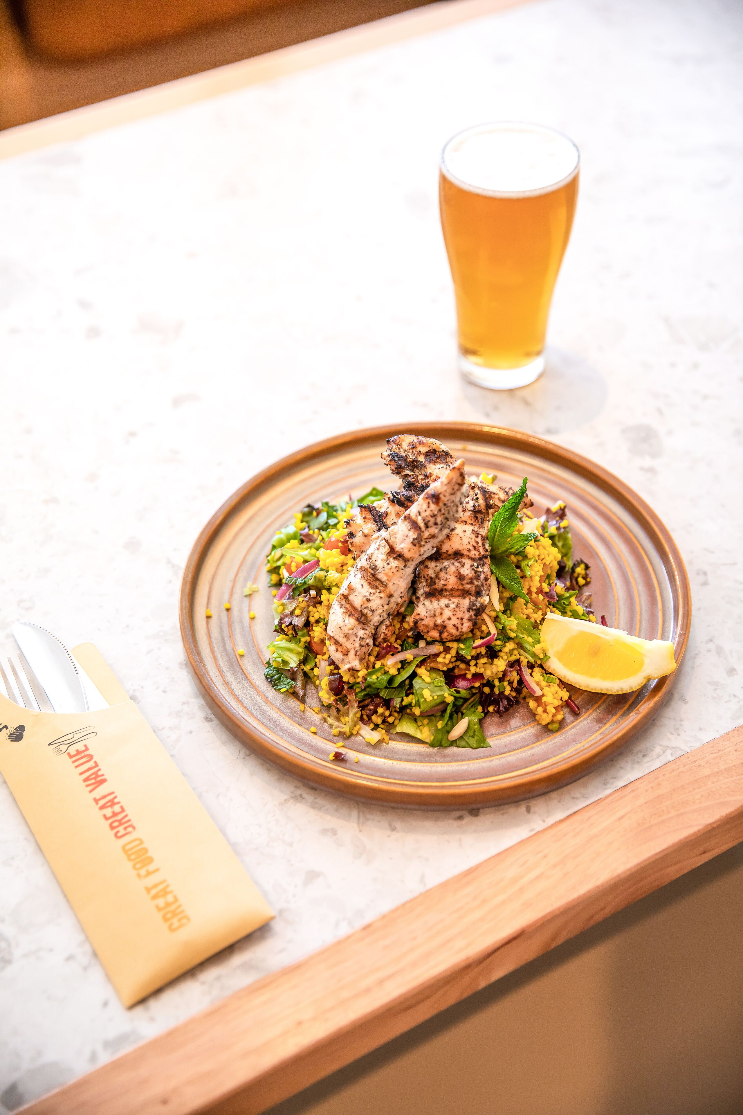 Un plato de pollo a la parrilla y ensalada de cuscús con un gajo de limón, acompañado de una cerveza sobre una mesa de mármol.