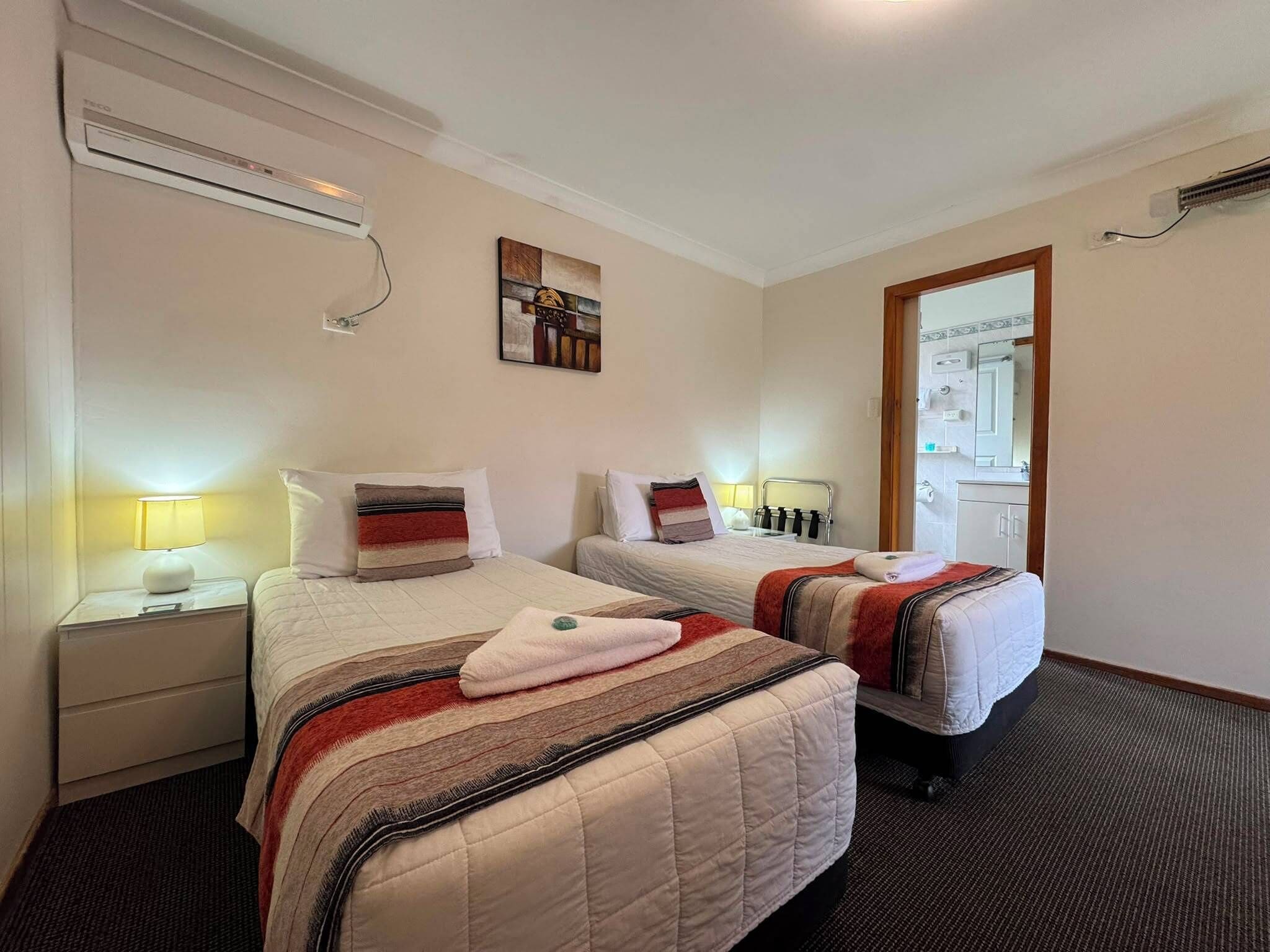 A hotel room with two beds, a nightstand with a lamp, a painting on the wall, and an air conditioning unit.