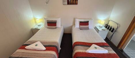 A hotel room with two beds, each with a pillow and a blanket, a bedside table with a lamp, a wall-mounted air conditioner, and a painting above the beds.
