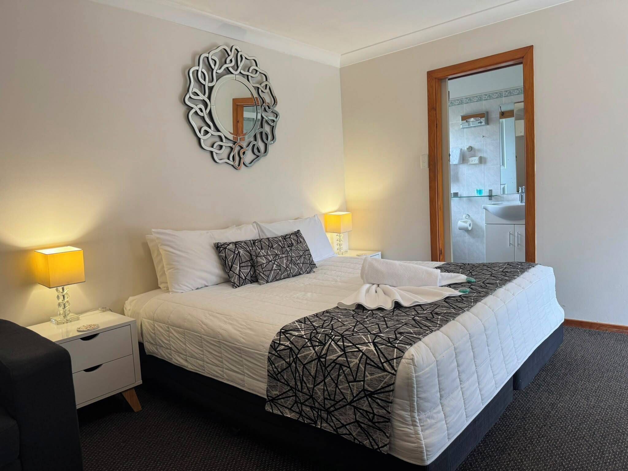 Un lit bien fait, recouvert d’une couvre-lit à motifs géométriques, deux lampes de chevet, un miroir décoratif et une porte en bois donnant sur une salle de bain.