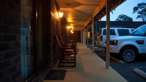 A covered outdoor area with a row of wooden benches, a brick wall, and a white SUV parked nearby.
