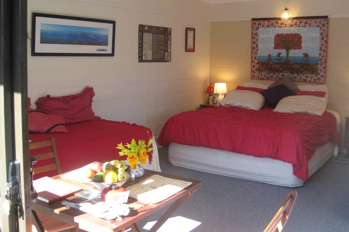 A room with two beds, a table with fruit and a vase, and a wall with a framed picture and a bulletin board.