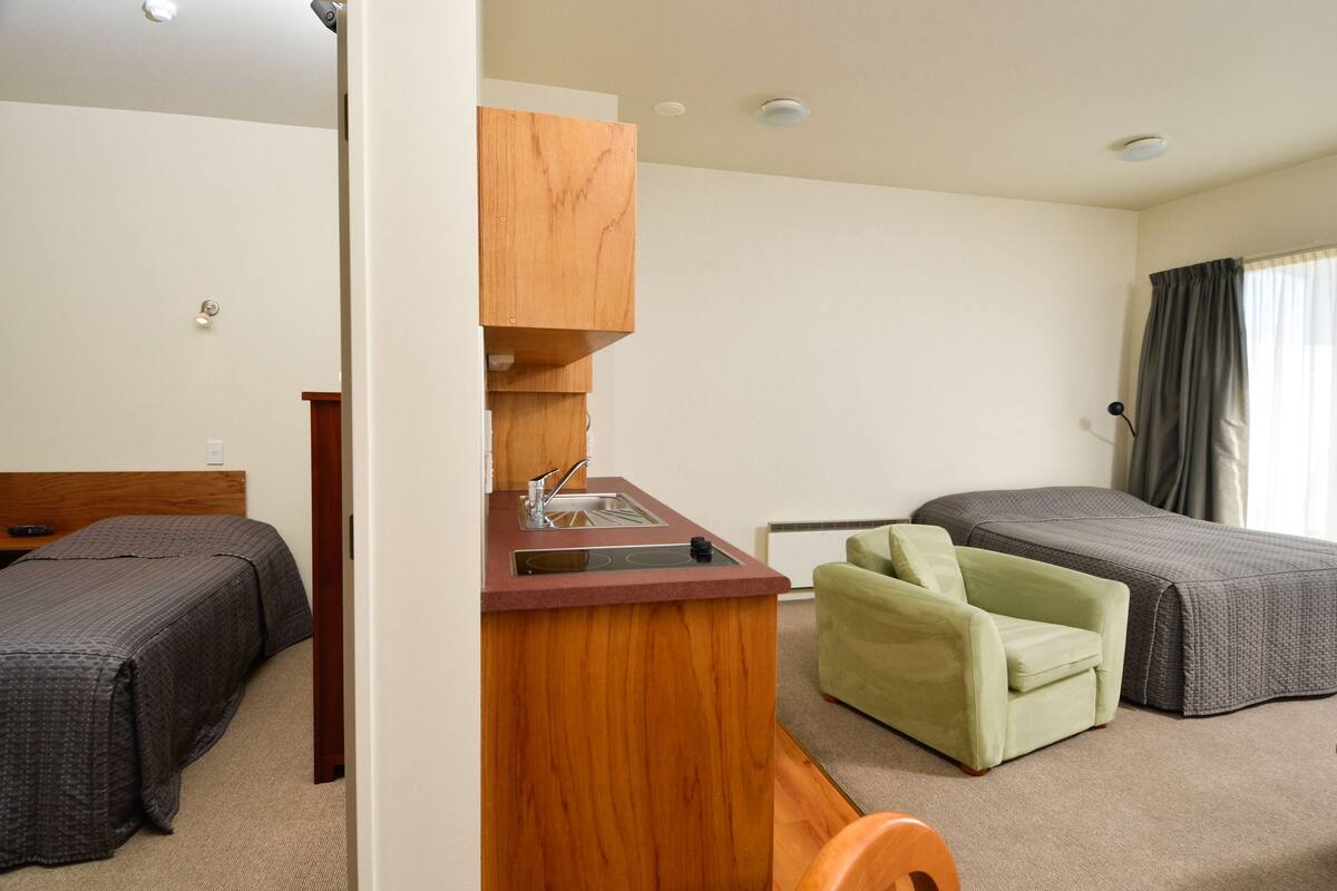 Habitación de hotel con dos camas individuales, una kitchenette pequeña, un sillón verde y una ventana con cortinas.
