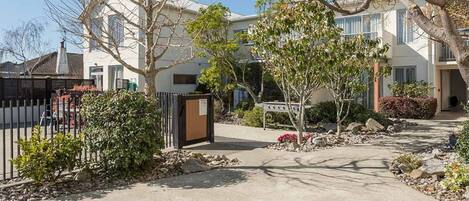 Un quartier résidentiel doté d’une entrée sécurisée, d’un chemin menant à un bâtiment à la façade blanche et d’un petit arbre au premier plan.