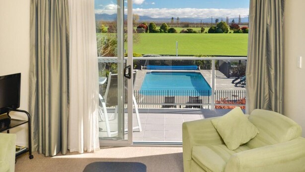 Una habitación con una puerta corrediza de vidrio que da a un balcón con piscina, césped verde y montañas al fondo.
