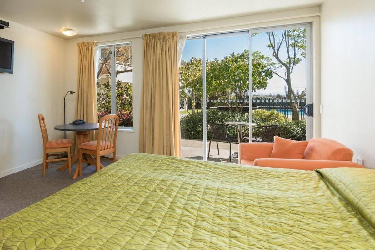 Habitación de hotel con cama, escritorio y silla. Se ve una piscina y árboles a través de la ventana.