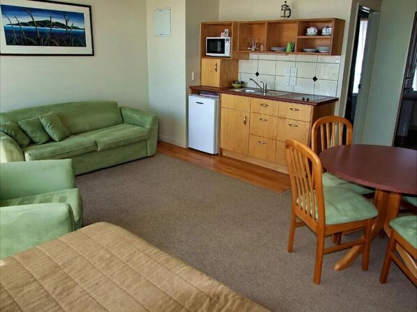 Un espacio habitable compacto con un sofá verde, una mesa de comedor con sillas, una zona de cocina con microondas y un refrigerador pequeño.