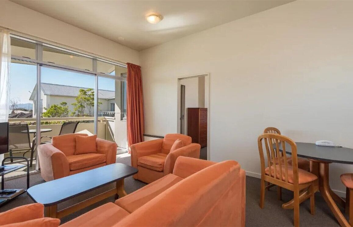 A room with a large window, orange sofas, a small table, and a wooden dining table with chairs.