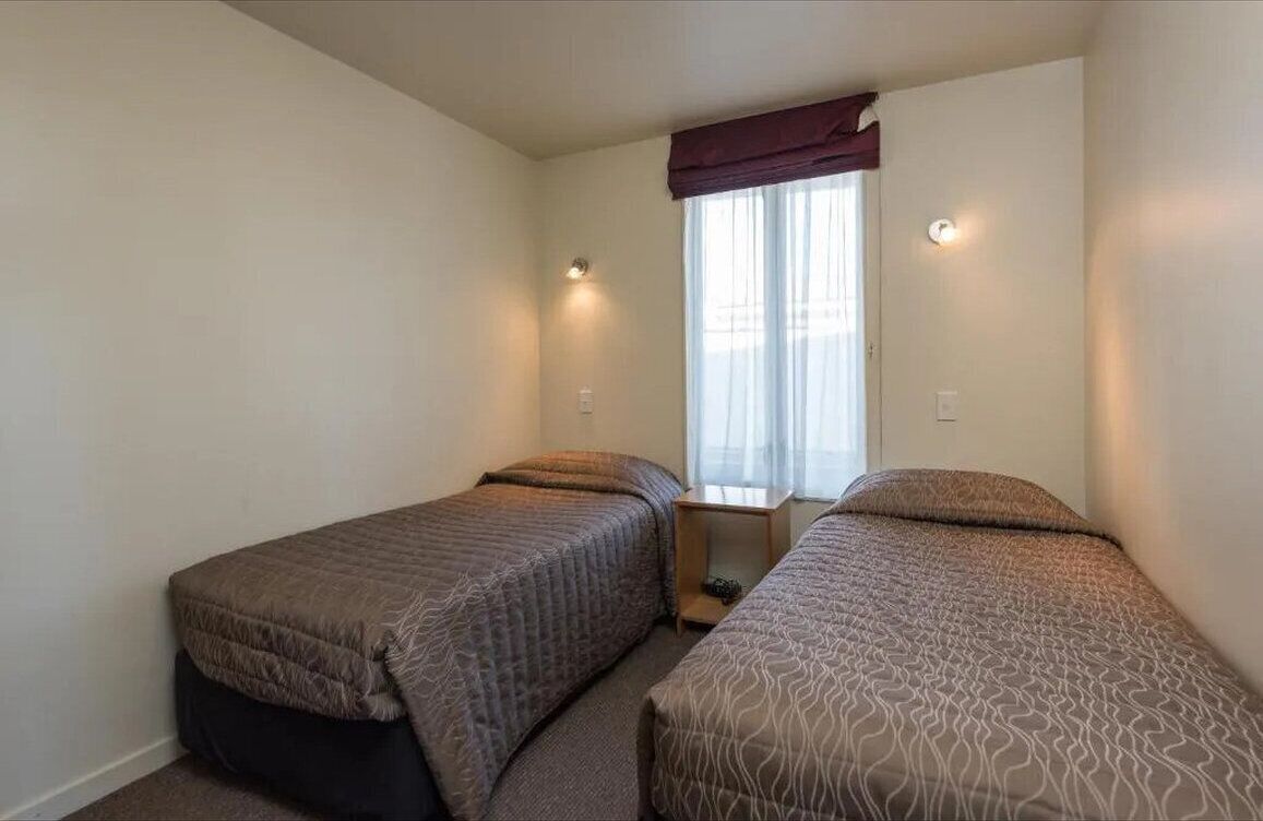 A small, single-room accommodation with two single beds, a window with curtains, and a nightstand between the beds.