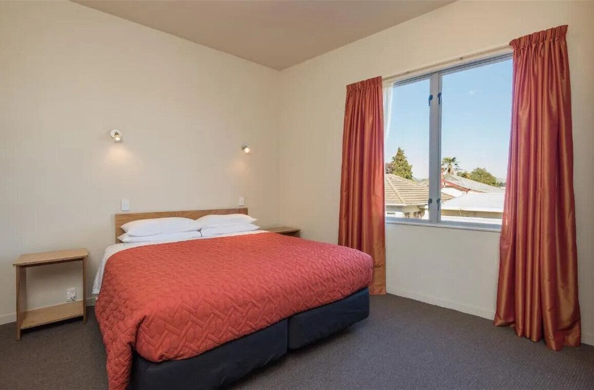 A bedroom with a bed, a nightstand, a window with curtains, and a view of buildings outside.